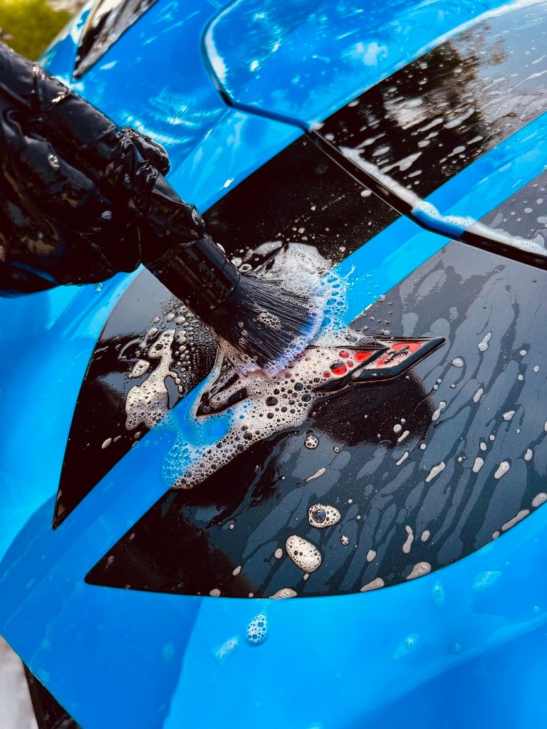 Soft brush cleaning the badge on a blue Corvette during a hand wash
